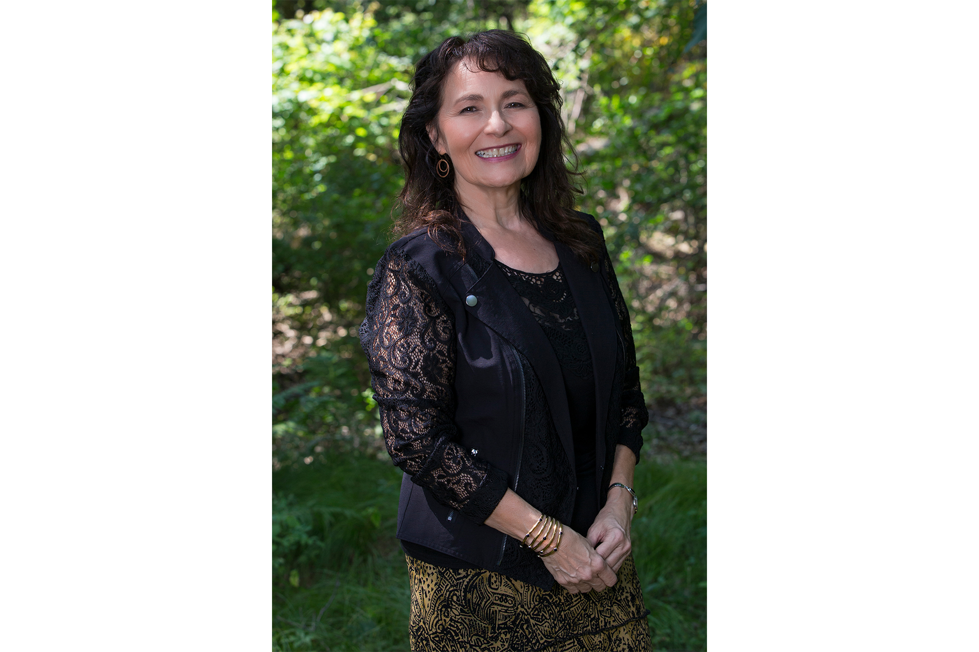 Portrait of Kimberly Blaeser, a light skinned woman in dark blue laced cardigan smiling in front of trees